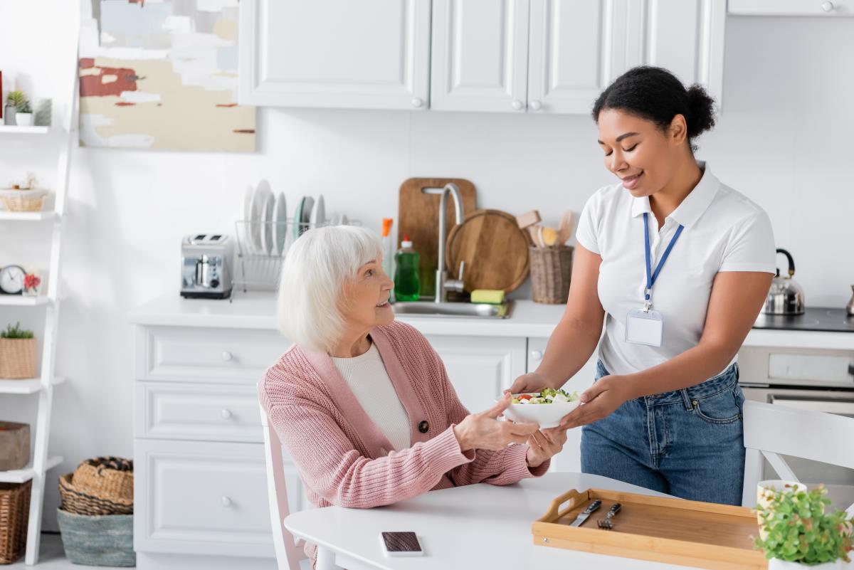 happy multiracial social worker holding bowl with salad for senior woman with grey hair